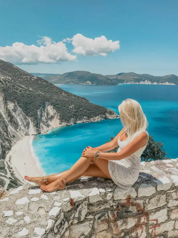 a woman sitting on a stone fence and looking at the sea